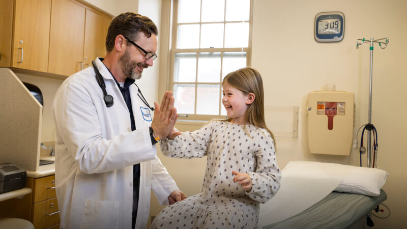 A man in a white doctor's coat with a stethoscope around his neck gives a high five to a young girl in an exam room.