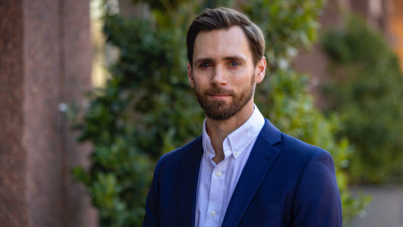 A man, Dr. Tyler Malone, is photographed from the shoulders up in front of some greenery. He has short hair, a beard, and is wearing a blazer and button down shirt. 