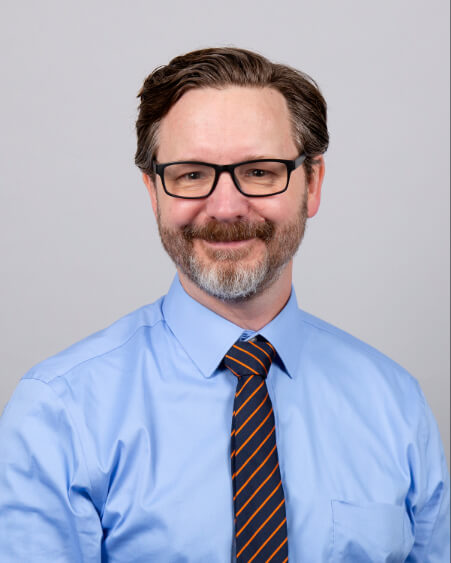 A man, Dr. Brandon Greene, is photographed. He is wearing a button down shirt, tie, and glasses.