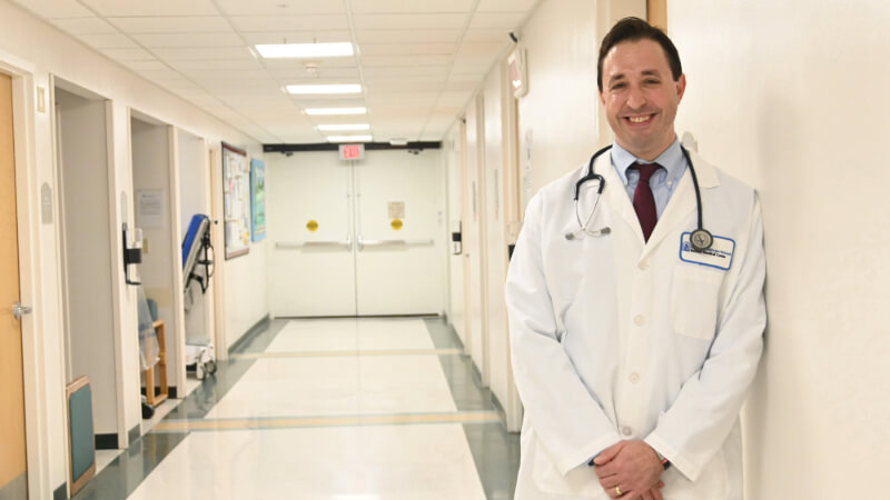 A doctor stands in a hospital hallway. He is smiling.