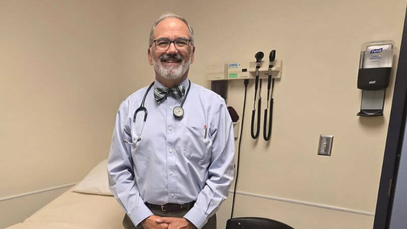 A man is standing in a medical exam room. He has a stethoscope around his neck. 