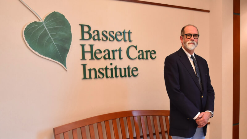 A man wearing a suit and tie is photographed in front of a sign that reads "Bassett Heart Care Institute"