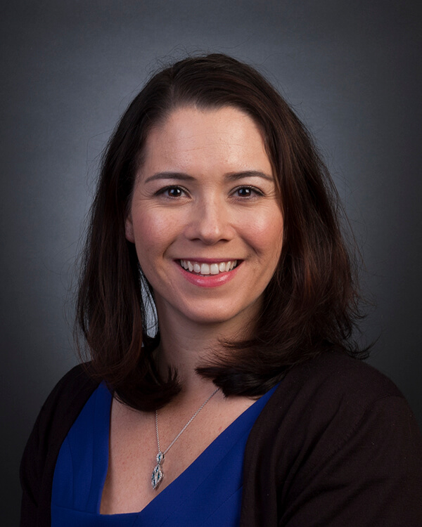 A woman is photographed from the shoulders up. She is wearing professional attire.