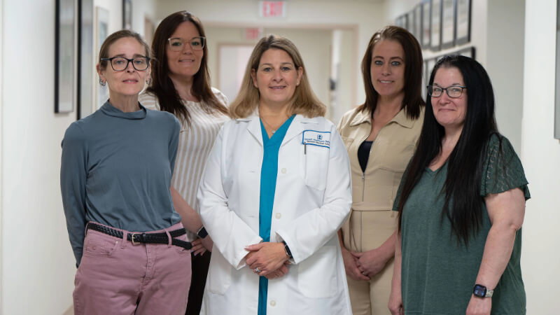 A group of five women stand in a hallway. The woman at the center is wearing a white doctor's coat and scrubs. The other women are wearing office attire. All five are smiling for the camera. 