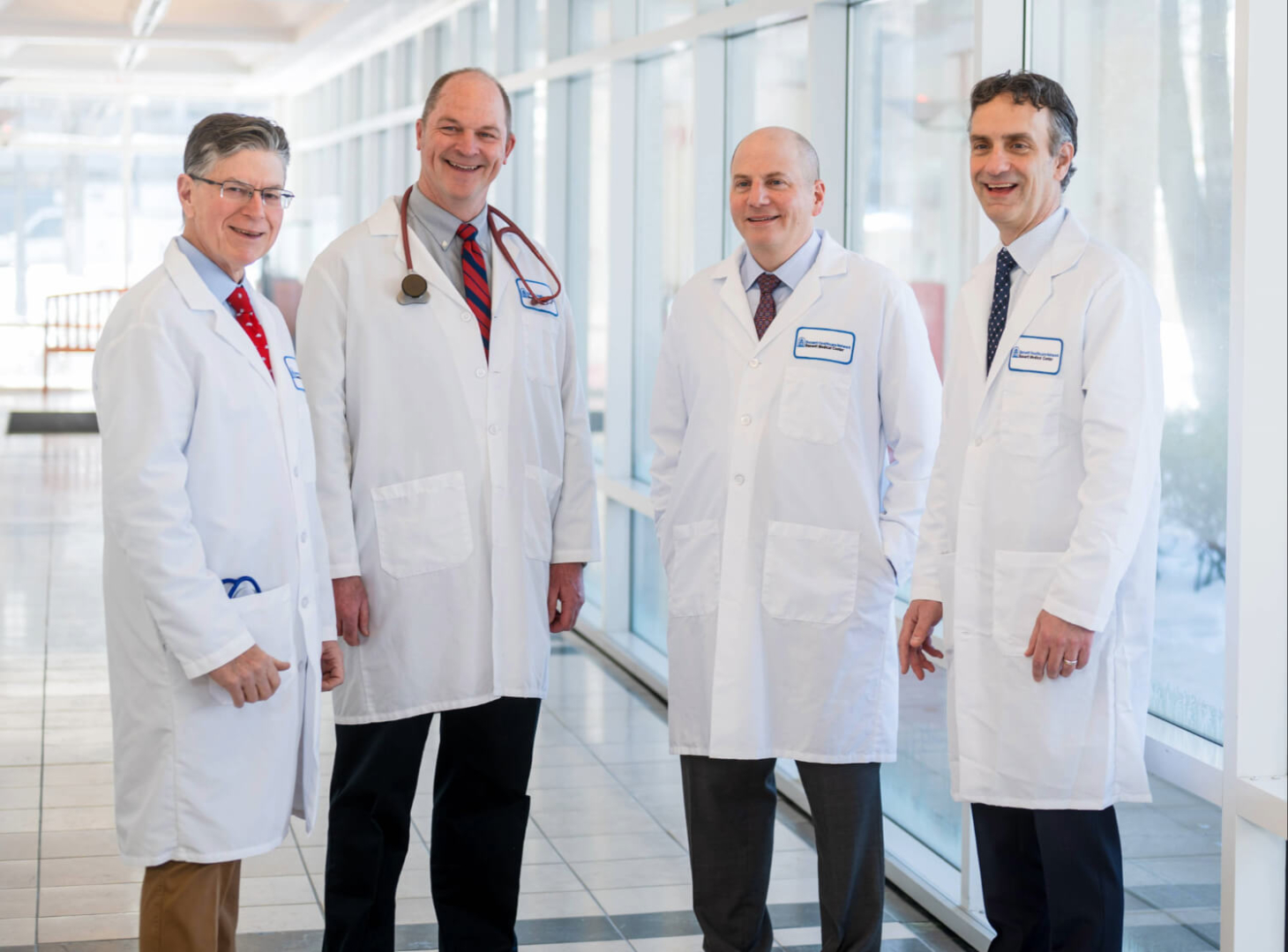 Four men, Dr. John Kelley, Dr. Michael Holmes, Dr. Evan Adelstein, and Dr. Anthony Nappi stand togther in a hallway. They are each wearing professional attire and white doctor's coats. 