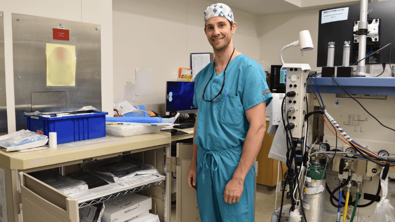 A surgeon in a cap and scrubs is photographed in an operating room. He is standing next to a storage cabinet. 