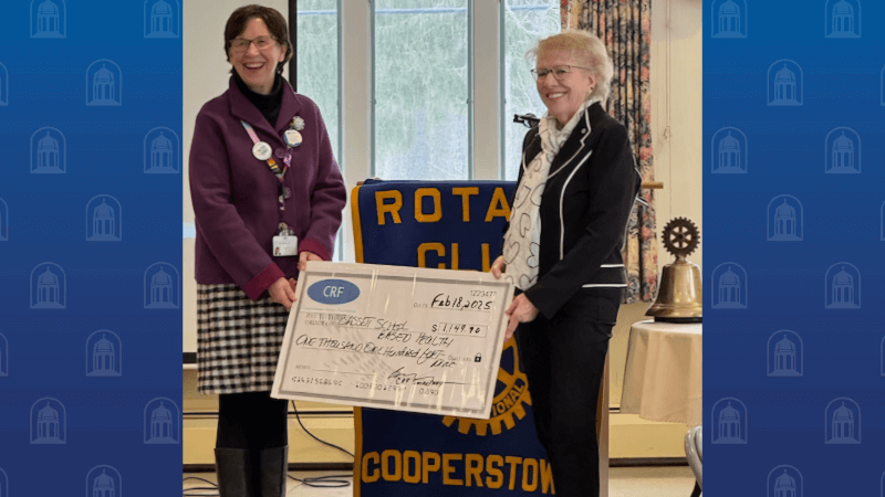 Two women hold up a ceremonial check in front of a podium. They are smiling. The podium behind them has a banner on it that appears to read Rotary Club Cooperstown.
